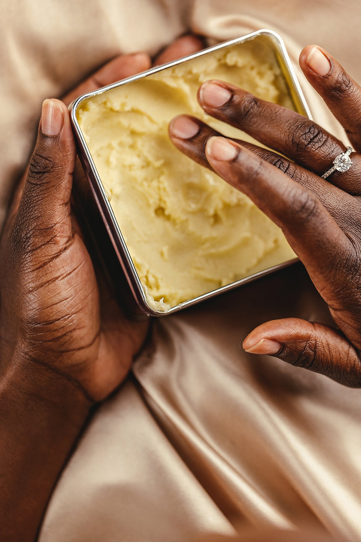Hand reaching into a large tin of shea butter to moisturize. on a soft background