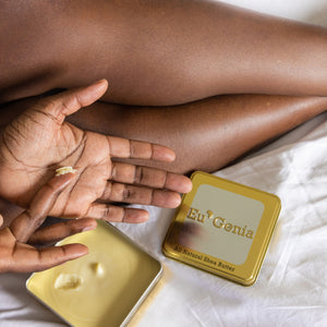 Person putting shea butter into the palm of their hand, open tin of shea butter in the background with bare legs visible.