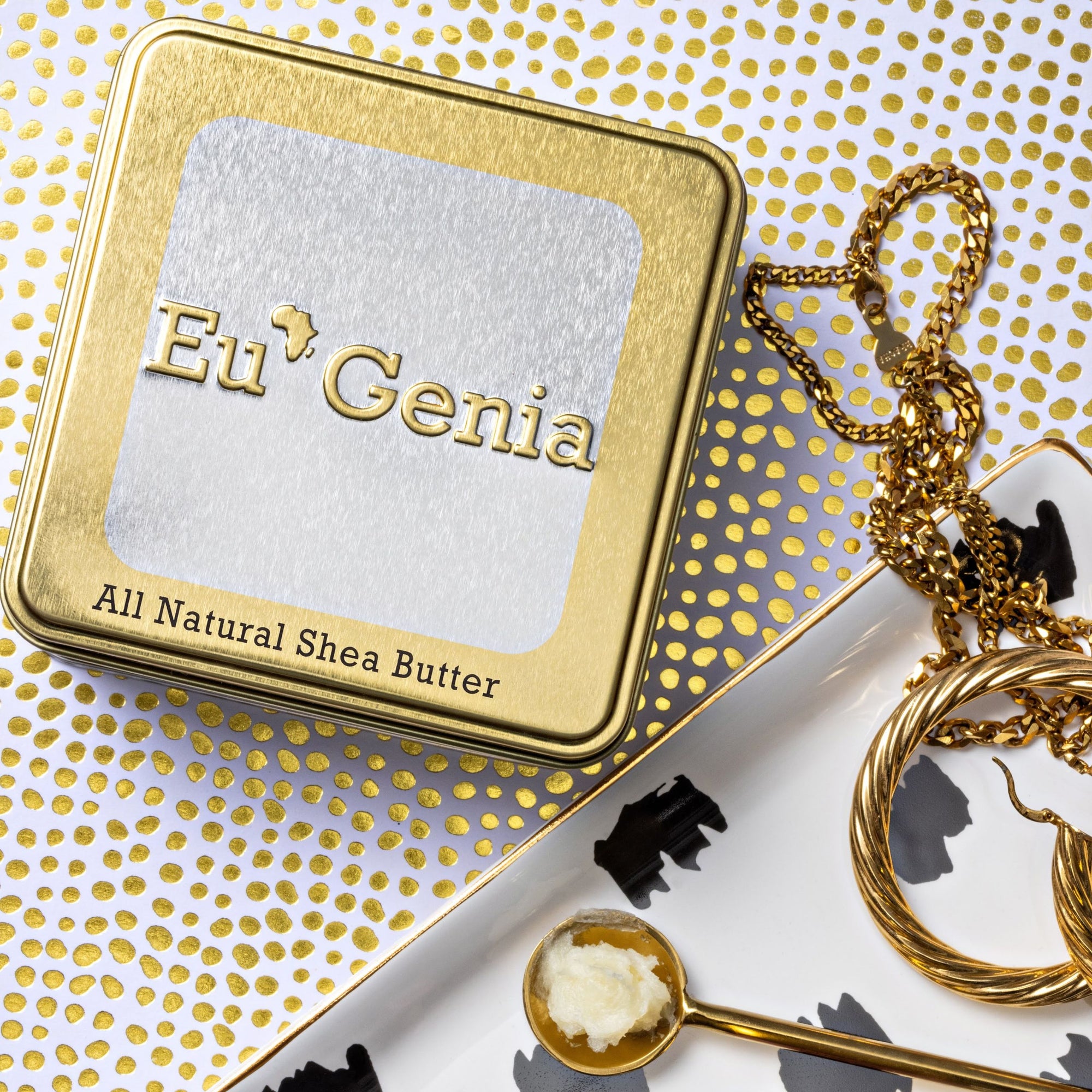 Gold Tin with text that reads "Eu'Genia All Natural Shea Butter". A white and gold background with a tray of gold jewelry and a gold spoon of shea butter sit next to the tin.