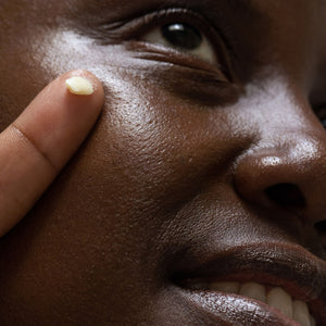 Close up image of a woman’s face with one finger holding shea butter and touching the side of their face.