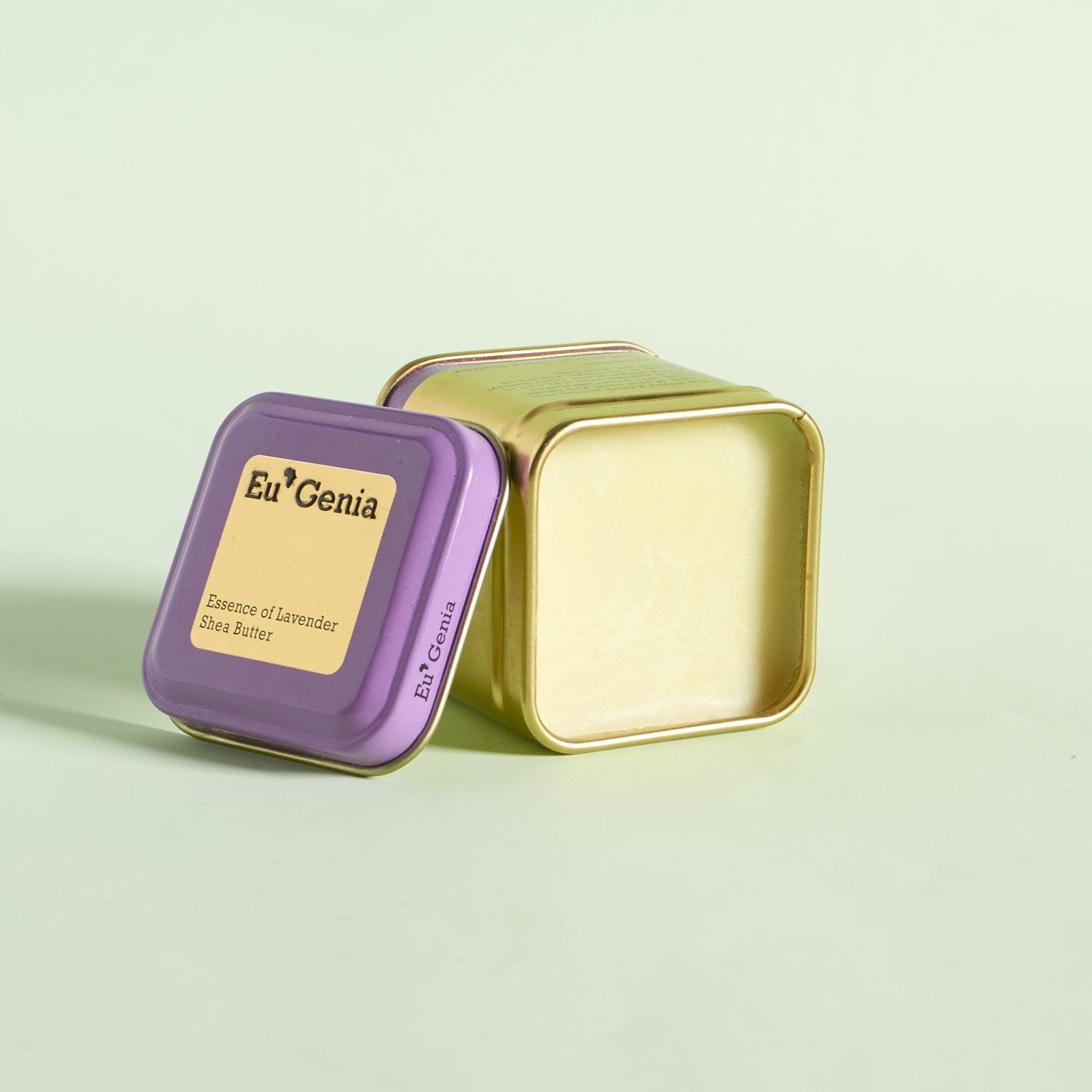 Gold tin filled with 100% natural shea butter and lid leaning against the tin that reads “Essence of Lavender”.